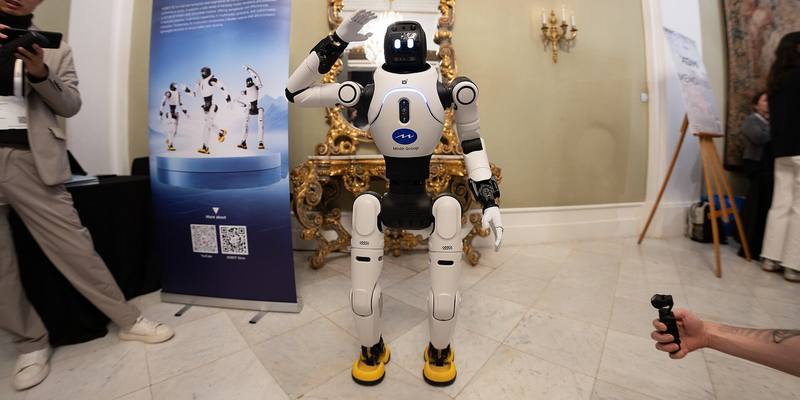 A white and black AgiBot X2 humanoid robot with yellow feet and a screen face raising one hand to wave, beside an AgiBot banner in an ornate room with a gilded mirror