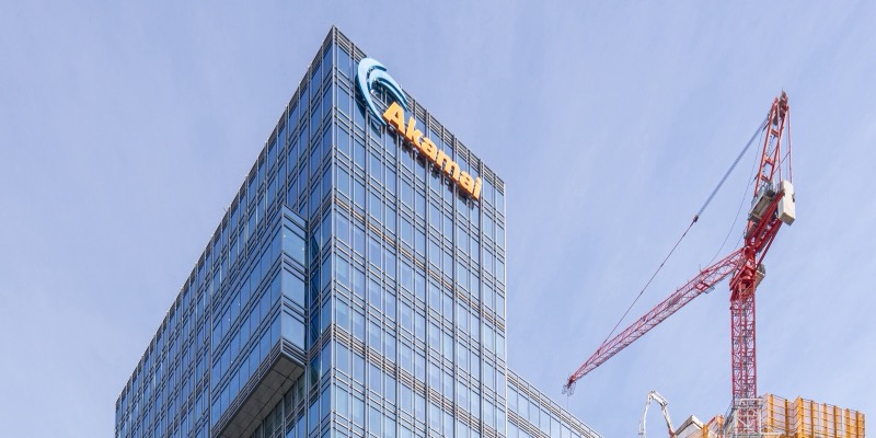 The top of Akamai’s glass office tower with the blue Akamai logo and orange lettering, next to a red construction crane against a blue sky