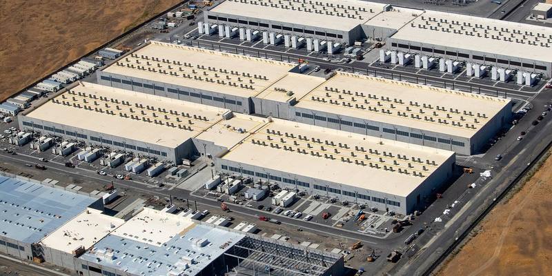 Aerial view of several large grey Amazon data centre buildings with rows of rooftop cooling units, surrounded by dry brown fields, with another building under construction in the foreground