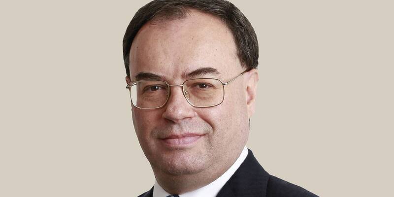 Andrew Bailey, in wire-rimmed glasses, a dark suit and a white shirt, smiling slightly in a head-and-shoulders portrait against a plain beige background.