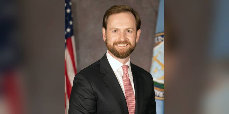 Andrew Ferguson smiling in a dark suit and red tie, with a US flag and the FTC flag behind him