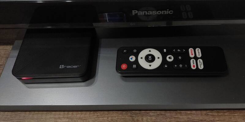 A small black Tracer Android TV box with a red standby light next to its remote control, on a grey stand beneath a television