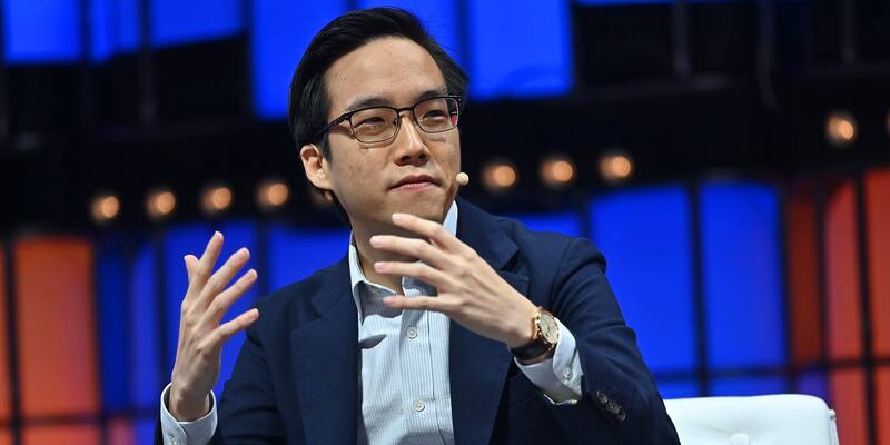 Andy Yen, in glasses, a navy blazer and a light blue shirt, gestures with both hands as he speaks on stage against a backdrop of blue and orange lights
