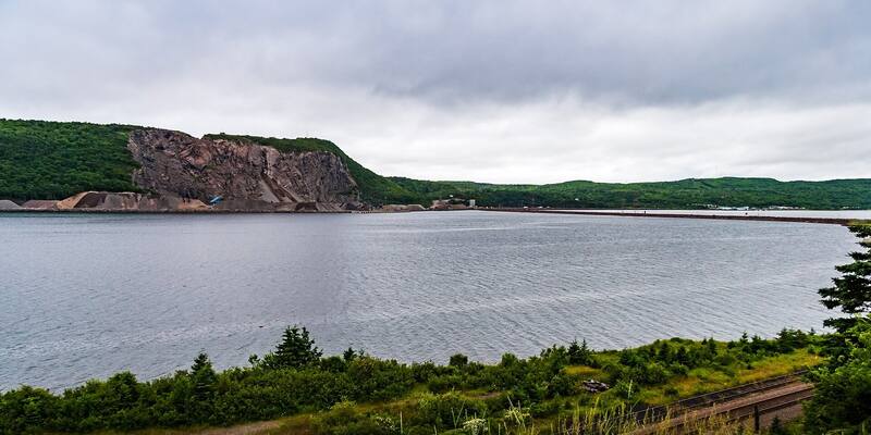 A wide grey strait under cloud, with a quarried rocky hillside and a long causeway on the far shore and green shrubs and a railway line in the foreground