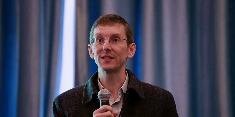 A man with short hair and glasses, wearing a dark jacket, speaking into a handheld microphone in front of blue curtains