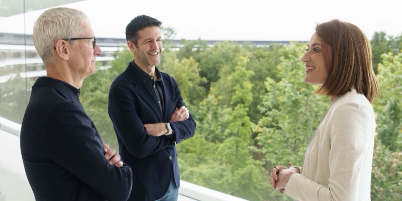 Tim Cook and John Ternus, arms folded, talking with Roberta Metsola in front of a window overlooking trees at Apple Park