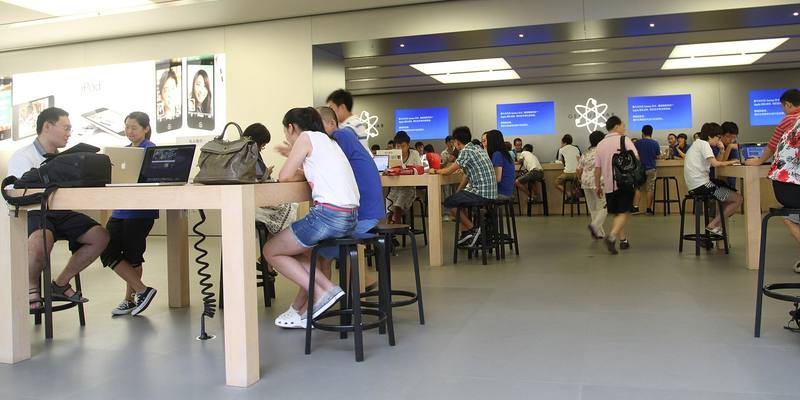 Customers sitting at wooden tables and a Genius Bar counter inside an Apple Store, with staff in blue shirts helping them