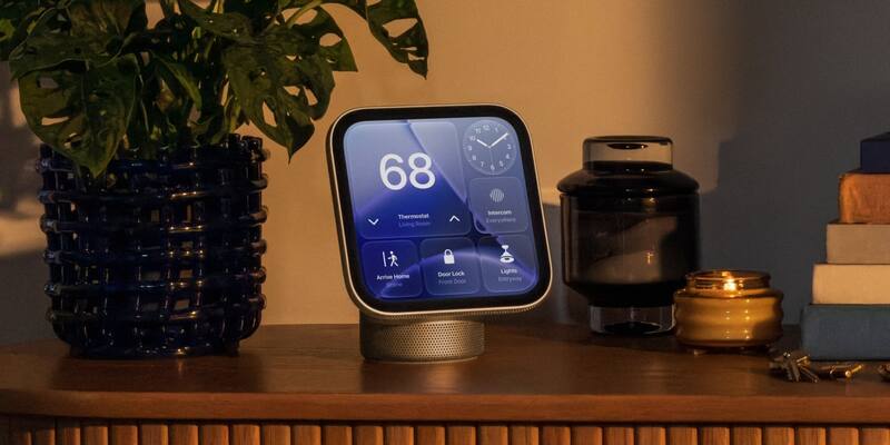 Apple HomeView smart display with a square screen showing thermostat, door lock and lights controls, on a wooden sideboard beside a plant and candles in warm evening light