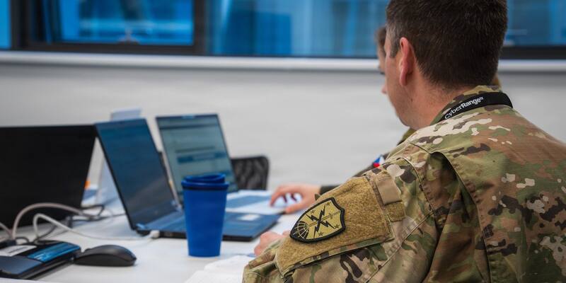 A US soldier in camouflage uniform with the Army Cyber branch shoulder patch sits at a table of laptops, seen from behind