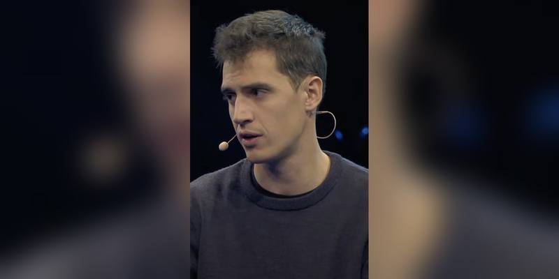 Arthur Mensch, wearing a dark jumper and a headset microphone, speaking on stage against a dark background.