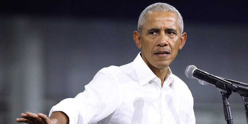 Barack Obama in a white shirt speaking at a microphone, one arm outstretched