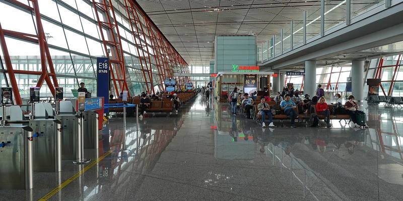 Travellers sitting in rows of seats beside boarding gate E18 in a long glass-walled airport terminal with red steel beams