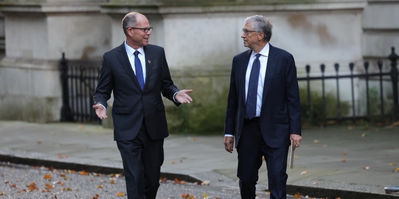 Bill Gates in a dark suit and blue tie walks along Downing Street beside Mark Suzman, who is talking and gesturing