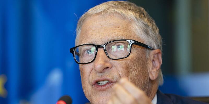 Bill Gates in black-rimmed glasses speaking and gesturing with one hand at a microphone, in front of a blurred blue EU flag
