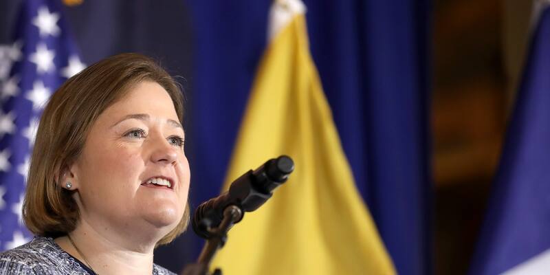 Brenna Bird, with shoulder-length brown hair and a blue tweed jacket, speaking into a microphone in front of US and Iowa flags