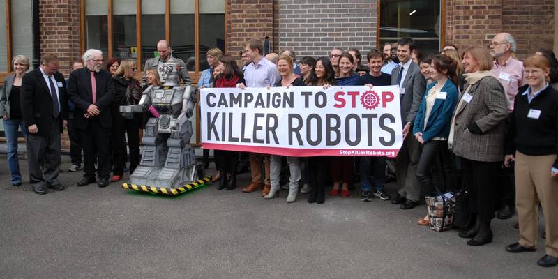 A large group of campaigners outside a brick building holding a banner reading Campaign to Stop Killer Robots, standing beside a person-sized grey toy robot