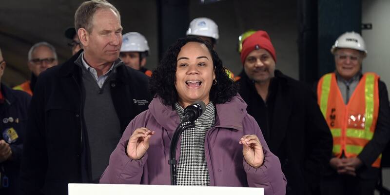 Carmen De La Rosa, with dark curly hair and a lilac coat, speaks at a microphone, with MTA officials and construction workers in hard hats standing behind her.