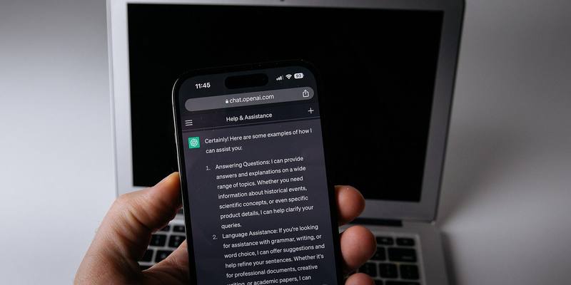 A hand holding a smartphone showing a ChatGPT conversation, with an open silver MacBook laptop behind it
