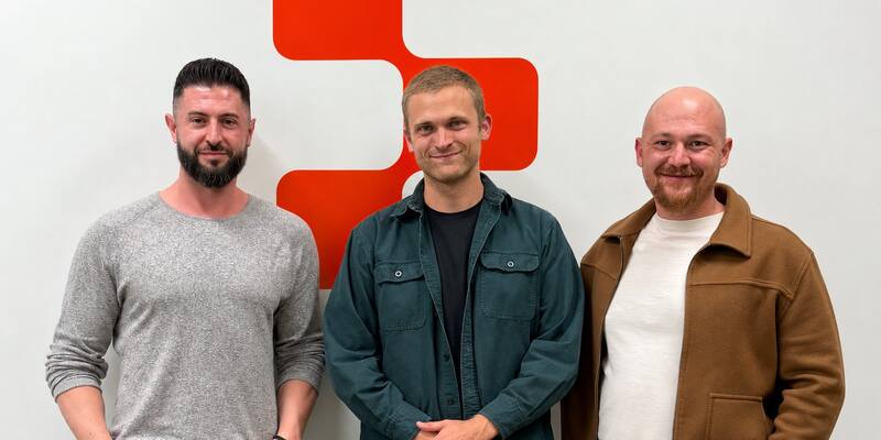 Three men stand smiling in front of a white wall with Replit’s orange logo: one in a grey jumper, one in a dark green shirt, one in a brown jacket