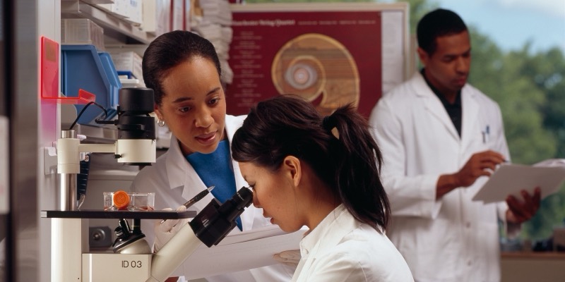 Three researchers in white lab coats in a laboratory, one looking into a microscope while a colleague watches