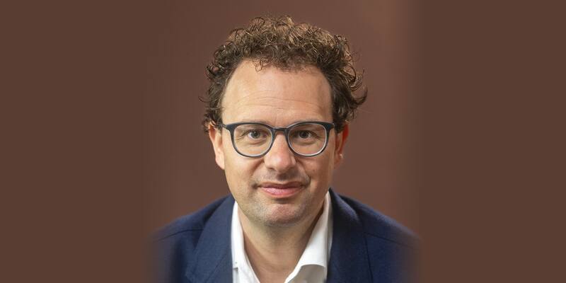 Dario Amodei, with curly hair and dark-rimmed glasses, wearing a navy jacket and white shirt against a brown background