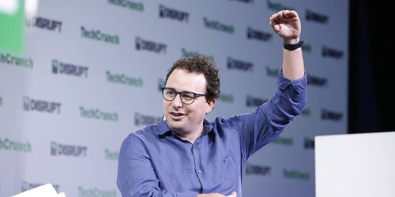 Dario Amodei, in glasses and a blue shirt, raising one arm as he speaks on stage in front of a TechCrunch Disrupt backdrop