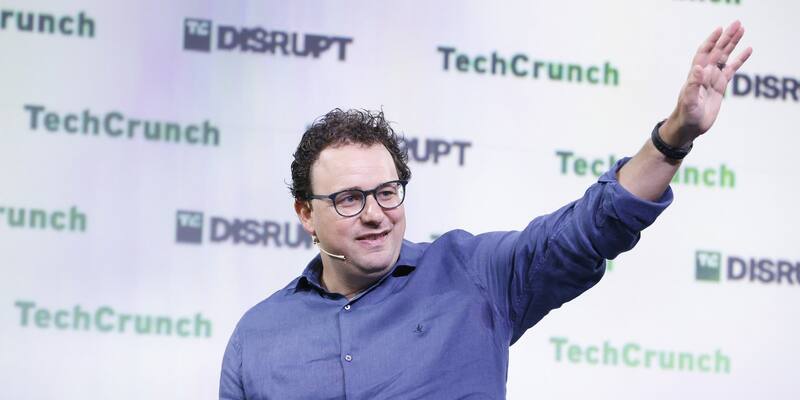 Dario Amodei, in glasses and a blue shirt, gesturing with one hand raised as he speaks in front of a TechCrunch Disrupt backdrop
