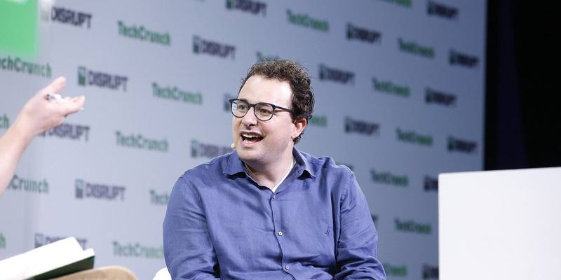Anthropic CEO Dario Amodei, in glasses and a blue shirt, speaking from an armchair on stage in front of a TechCrunch Disrupt backdrop