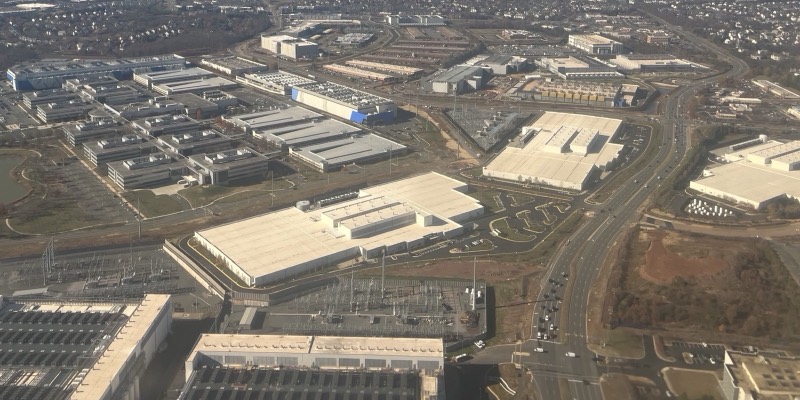 An aerial view of large white data centre buildings and electrical substations beside roads, with housing estates in the background
