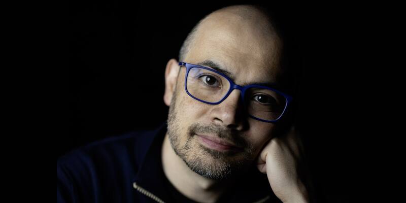 Demis Hassabis, bald with a short grey-flecked beard and blue-framed glasses, resting his chin on his hand against a black background