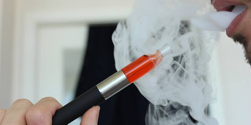 Close-up of a hand holding a black and silver vape pen with orange liquid while a man's mouth breathes out a thick white cloud of vapour