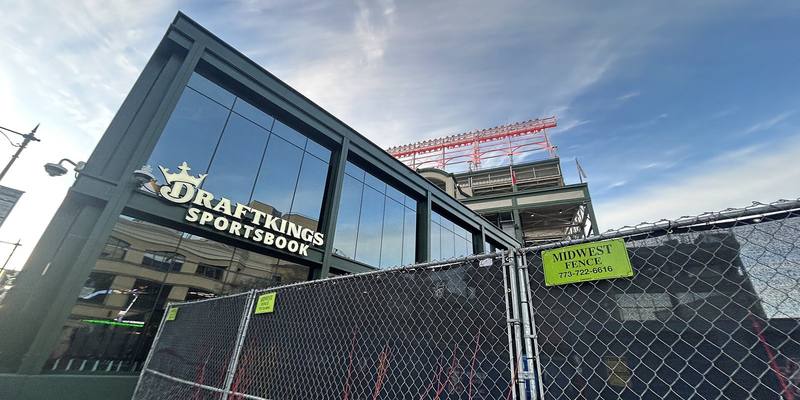 A glass-fronted building with a DraftKings Sportsbook sign beside Wrigley Field, behind a chain-link fence under a blue sky.