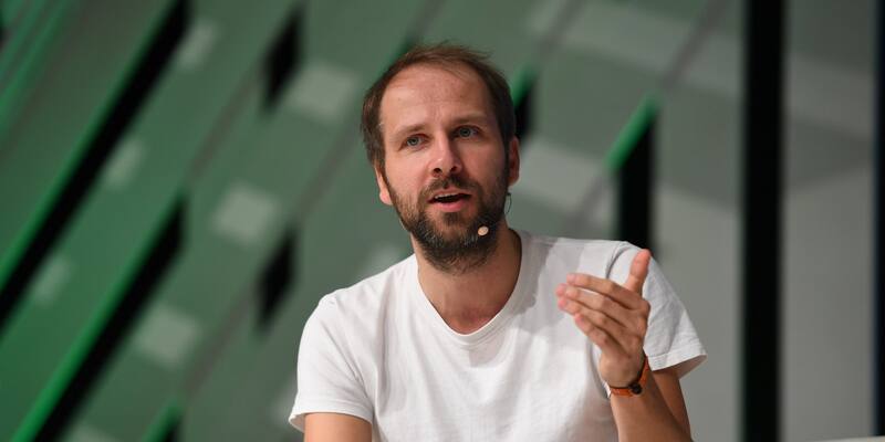 Christian Kroll, a bearded man in a white T-shirt with a headset microphone, gestures as he speaks on stage in front of a green and grey backdrop