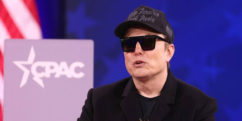 Elon Musk in a black cap, sunglasses and black jacket speaking on stage in front of a CPAC sign and a US flag.