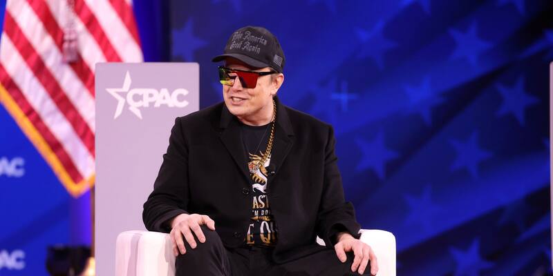 Elon Musk in a black cap and tinted sunglasses, seated in a white chair on stage in front of a CPAC sign and a US flag.