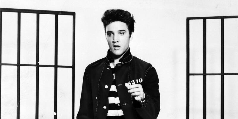 A young Elvis Presley in a black prison-style jacket with the number 6240 over a striped shirt, pointing, between two sets of jail bars