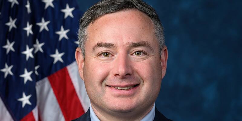 Eugene Vindman, smiling, in a dark suit, light blue shirt and striped red and blue tie, in front of a US flag and a dark blue backdrop