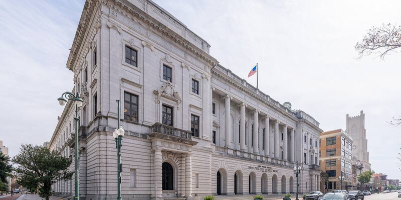 The white marble John Minor Wisdom US Court of Appeals Building in New Orleans, with columns, arched ground-floor windows and a US flag on the roof, under a pale sky
