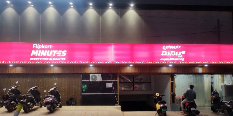 A shopfront at night with a bright pink Flipkart Minutes sign in English and Kannada, and scooters parked outside