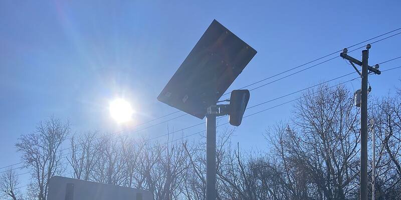 A Flock Safety licence plate reader camera with a solar panel on a pole beside a road, against a bright blue sky with bare trees