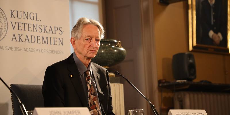 Geoffrey Hinton in a dark suit and patterned tie sitting at a press conference table with microphones, in front of a Royal Swedish Academy of Sciences banner