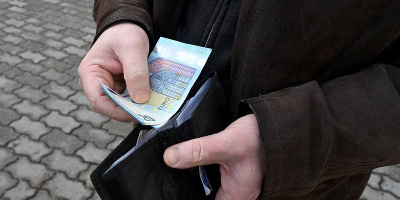 Close-up of a man's hands in a brown leather jacket opening a black wallet and taking out a folded 20 euro note, with grey paving stones behind