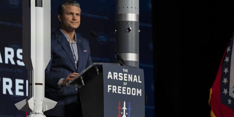 Pete Hegseth in a navy jacket at a podium reading The Arsenal of Freedom, standing between two missile models in front of a dark blue backdrop
