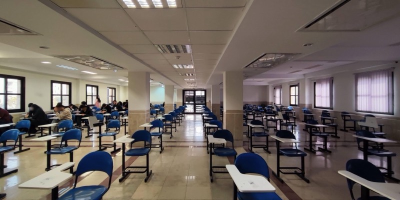 A university exam hall with rows of blue chairs and desks, students sitting an exam on the left