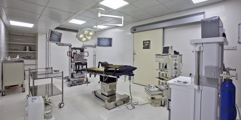 An empty, brightly lit hospital operating theatre with a surgical table under a ceiling lamp, two monitors on arms, an anaesthesia machine, trolleys and a blue gas cylinder