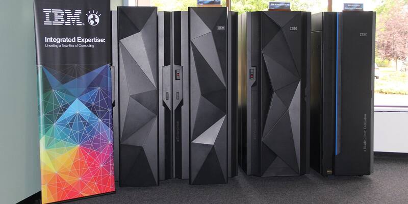 A row of tall black IBM zEnterprise mainframe cabinets with angular faceted doors, beside a colourful IBM banner, in a bright room with windows.
