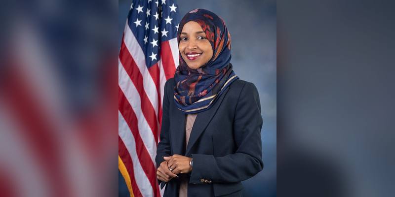 Ilhan Omar smiling in a patterned navy headscarf and dark blazer, standing in front of a US flag