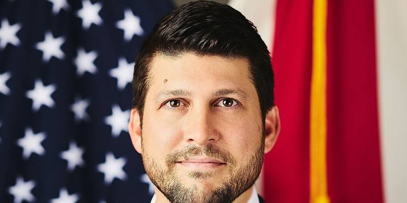 Close-up of James Uthmeier, a bearded man with dark hair, in front of the US and Florida flags