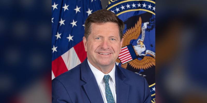 Jay Clayton smiling in a blue suit and patterned tie, in front of a US flag and the seal of the Director of National Intelligence