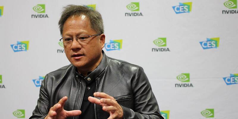 Jensen Huang in glasses and a black leather jacket gesturing with both hands in front of a white wall printed with Nvidia and CES logos